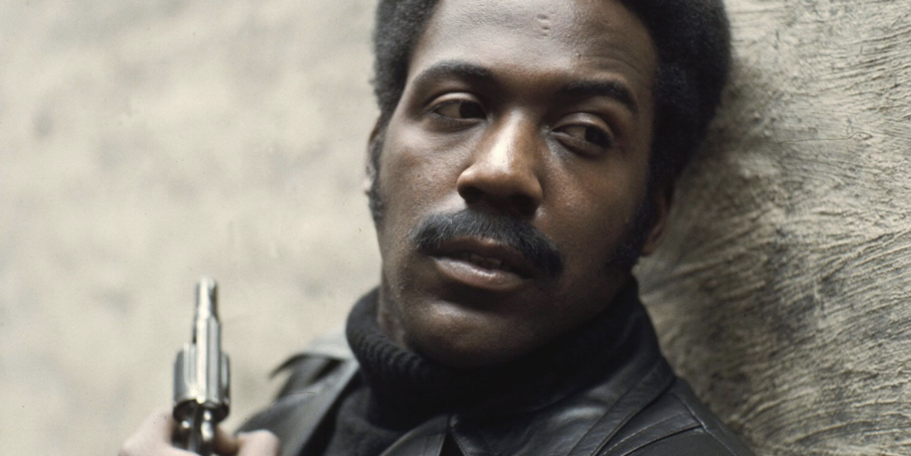 Richarad Roundtree, playing Shaft, stands with his back against a wall wearing a dark turtleneck sweater and leather coat. We see a close-up of his face.