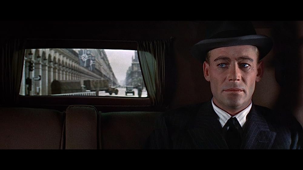 Sitting on the right inside a dark car is Peter O'Toole's character, General Tanz. He is wearing a black suit and a black bowler hat. To his left is a window showing a Nazi-occupied city.