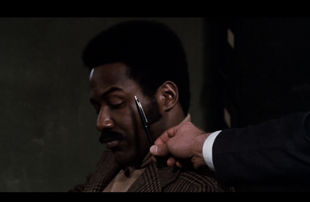 Shaft looks down as a black pen is held up next to his face by a white hand. 