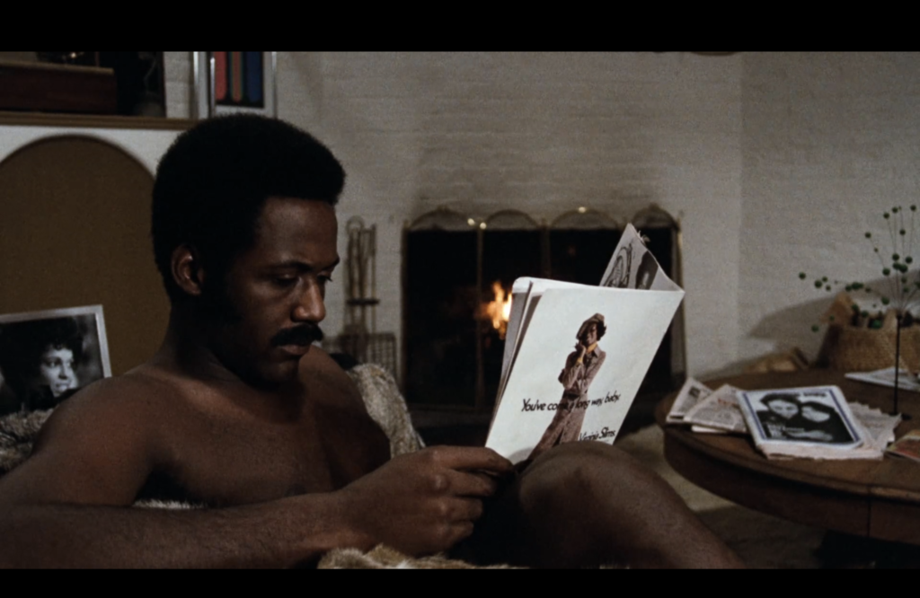 Shaft reads a magazine shirtless on the couch, with a roaring fire in the background. 