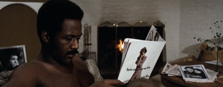 Shaft reads a magazine shirtless on the couch, with a roaring fire in the background.