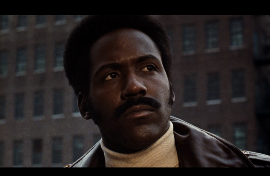 Shaft looks into the distance, wearing a beige turtleneck and brown coat. 
