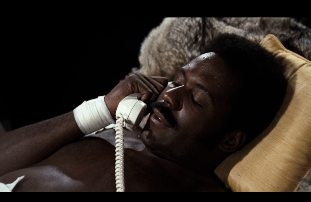 Shaft lies on a couch, holding a white telephone to his ear. 