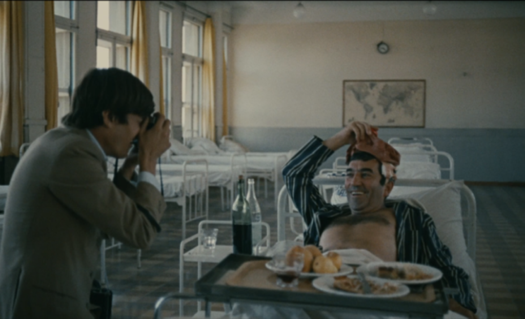 On the left is a man taking a photo, on the right is a man laying in a hospital bed with a tray of food in front of him. He is smiling and holding an ice pack on his head.