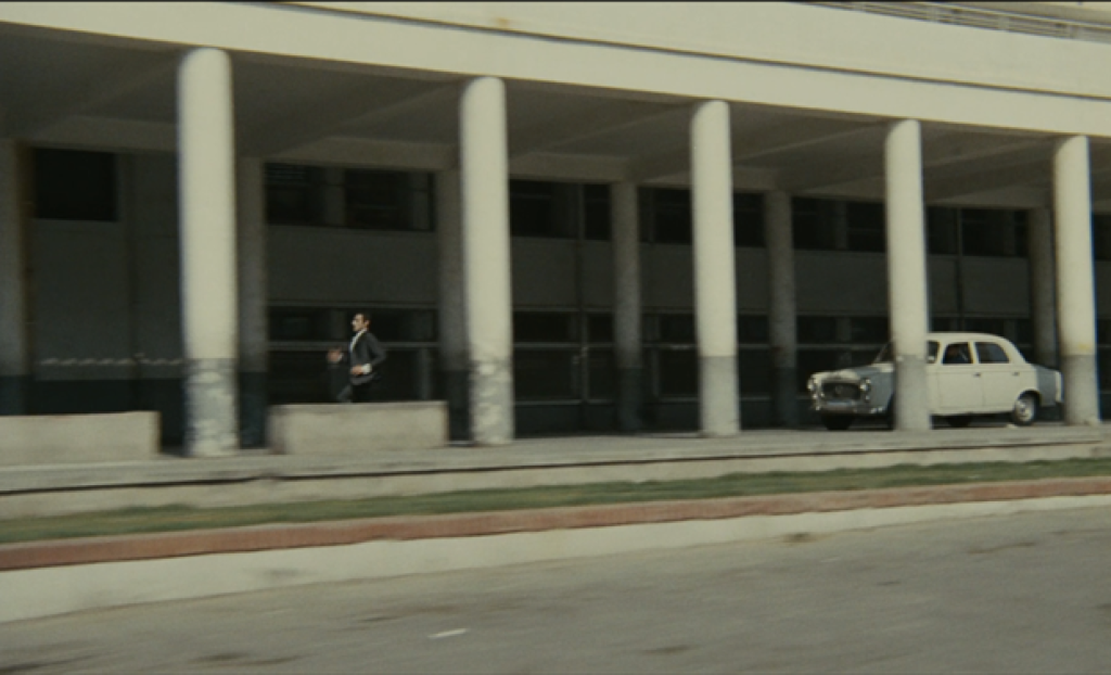 A man is running under a white concrete building, with a car following him.