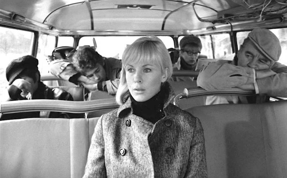 A white, blonde woman sits in the middle of a bus with one woman and one man seated on either side behind her.