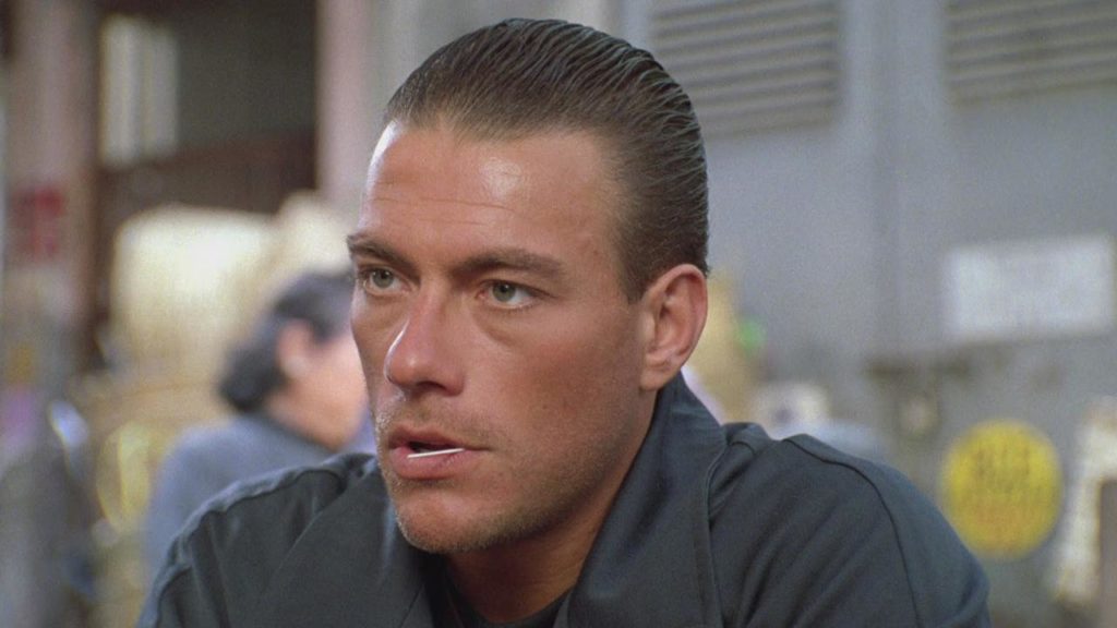 A close-up of Jean-Claude Van Damme as Alex, scruffy and serious with a toothpick in his mouth against a blurred background.
