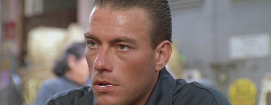 A close-up of Jean-Claude Van Damme as Alex, scruffy and serious with a toothpick in his mouth against a blurred background.