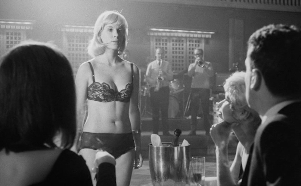 A white, blonde woman wearing a black bra and panties stands in front of a table of three people seated with their backs to the viewer.