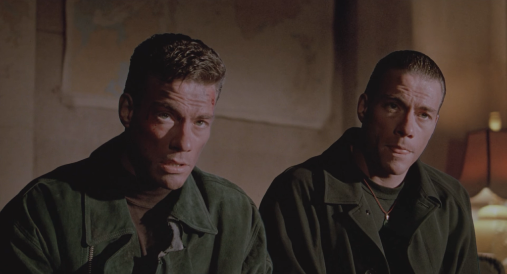 A medium two-shot of Jean-Claude Van Damme as both Chad, with wavy hair, and Alex, with slicked back hair, both in dark jackets against a poorly double exposed, dim background.