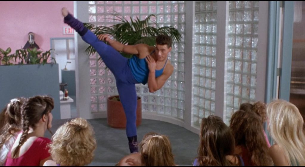 A long shot of Jean-Claude Van Damme in brightly colored spandex as Chad kicking high in the air with several women students watching, against a background of pastel paint and a gloriously 80s glass block half-wall.