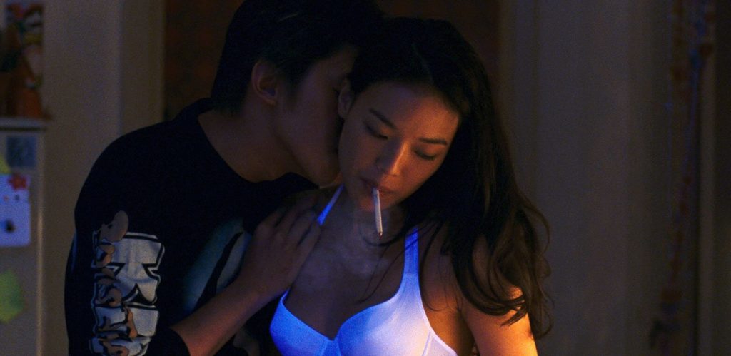A woman named Vicky, played by actress Shu Qi, is standing with her lover Hao-Hao, played by Duan Chun-hao, leaning onto her, placing a hand on her shoulder and kissing her neck. The woman is wearing a white bra that is bathed in blue light and smoking a cigarette while looking disinterested in the man's advances. The man is wearing a black longsleeve shirt