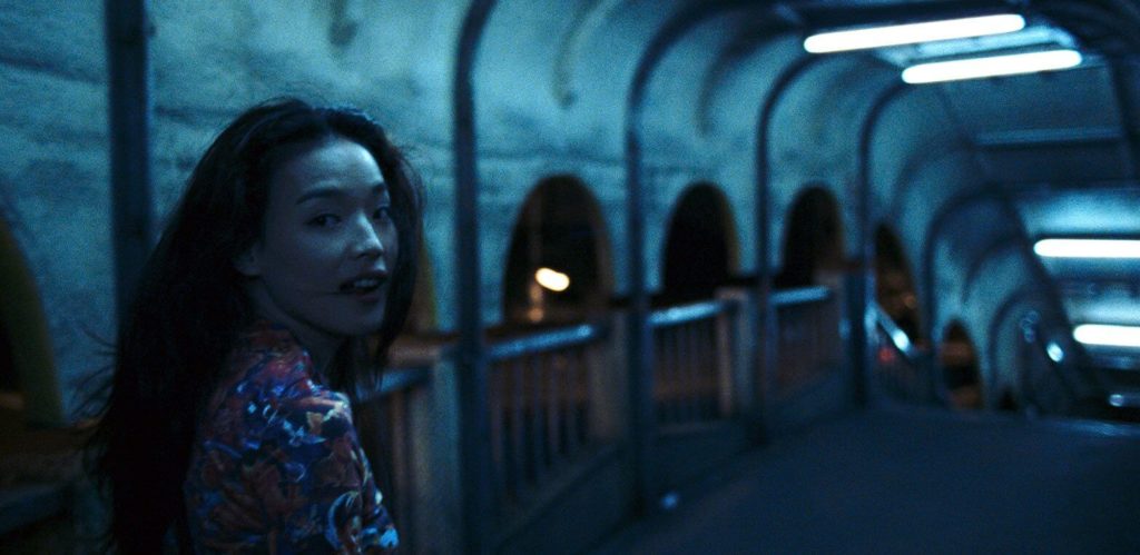 A woman named Vicky, played by actress Shu Qi, is walking through a pedestrian overpass at night, with the lighting putting a blue hue on her and her surroundings. She is looking back over her shoulder at the camera.