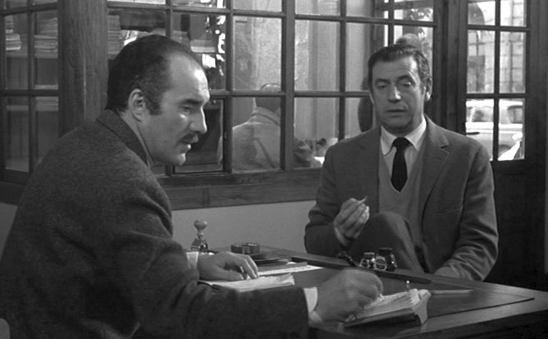 In a police station, a mustached man sits on the left side of the table, interrogating a handsome stranger on the right.