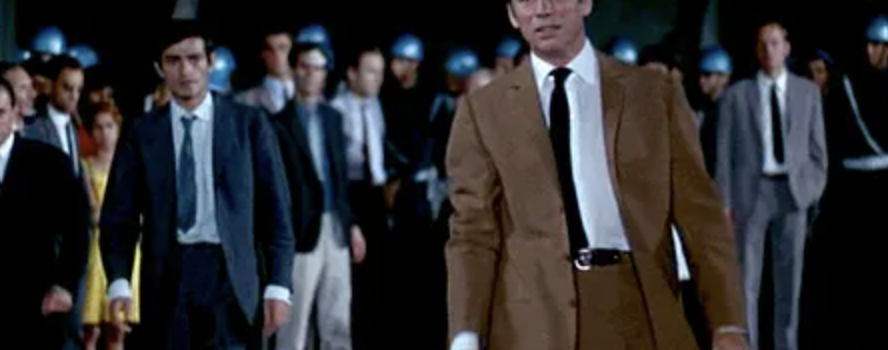 A crowd of men stand on the road at night; in the back is a line of men wearing blue helmets. In front of them are mostly men in suits. At the front is Yves Montand, dressed in a brown suit.