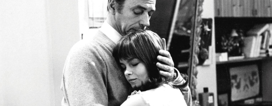 In an empty kitchen, a tall man in a soft sweater embraces a shorter woman with a bob haircut.