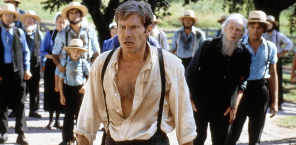 Hot, hunky Harrison Ford stands in front of a crowd of Amish people, wearing an open cream button down and black suspenders.