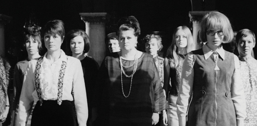 A crowd of women dressed in 60s era clothing stare at an unseen figure off camera.
