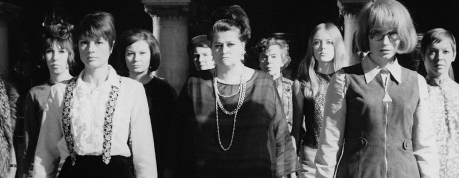 A crowd of women dressed in 60s era clothing stare at an unseen figure off camera.