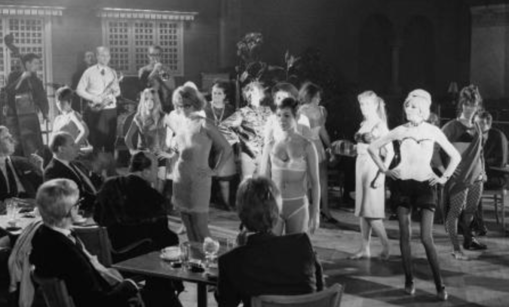 Two tables of men sit in front of a room filled with women who are wearing underwear.