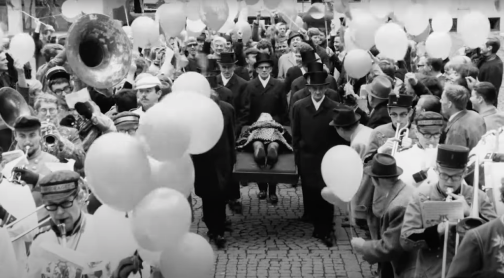 A procession of men in black carry a body through a marching band.