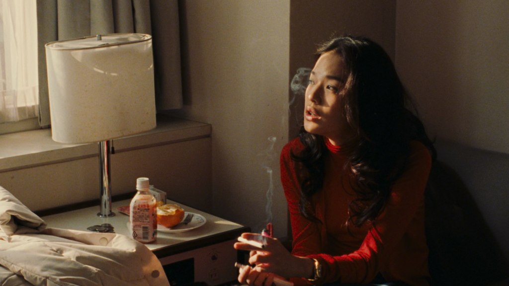 A woman named Vicky, played by actress Shu Qi, sits beside an empty bed in an apartment and gazes out the window, where sunlight is spilling in. She is wearing a red dress and holding a lit cigarette between her fingers. On the bedside table next to her, there is a lamp, a half-drunk bottle of juice, and a half-peeled and eaten orange on a plate with a fork