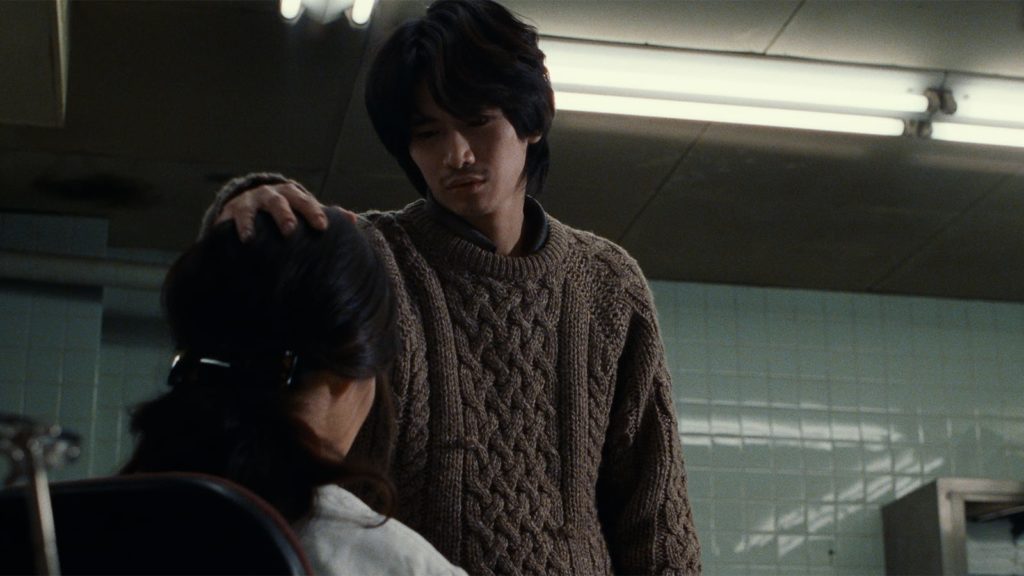 A young asian man with light skin and dark hair of medium length stands in a mint-tiled room facing the camera, placing his hand on a dark-haired woman's head who is seated facing away from the camera. 