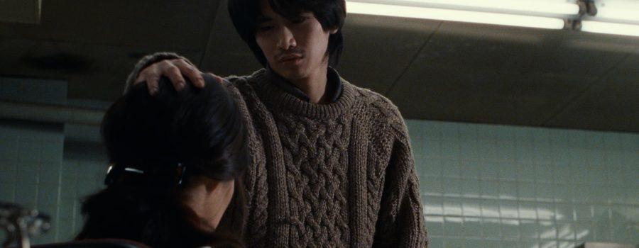 A young asian man with light skin and dark hair of medium length stands in a mint-tiled room facing the camera, placing his hand on a dark-haired woman's head who is seated facing away from the camera.