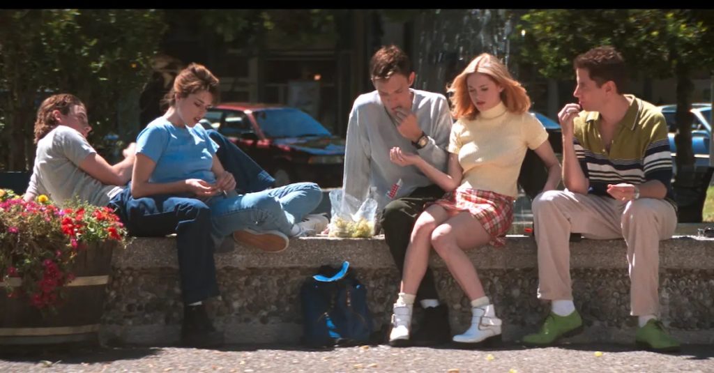 Five white teenagers (some of the main cast of the movie) are sitting by a water fountain on a sunny day