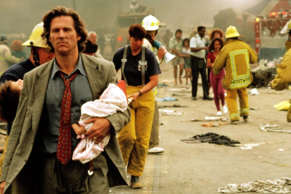 Max Klein (Jeff Bridges), a white adult man with medium long brown hair wearing a grey-green suit, grey-blue shirt, and red patterned tie, walks way from a scene (and toward the camera) of destruction holding a baby wrapped in a blanket. Firefighters, emergency vehicles, and other survivors populate the background.