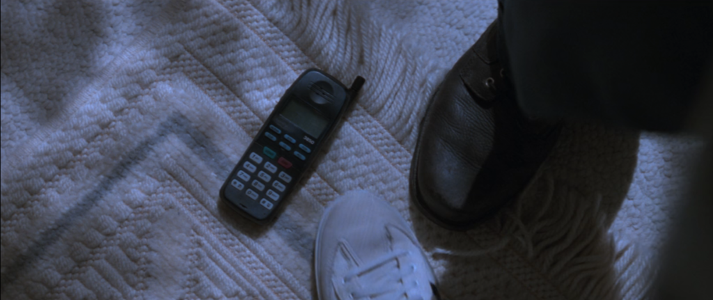 A 90s cell phone rests on the floor between the feet of Sidney Prescott and Billy Loomis