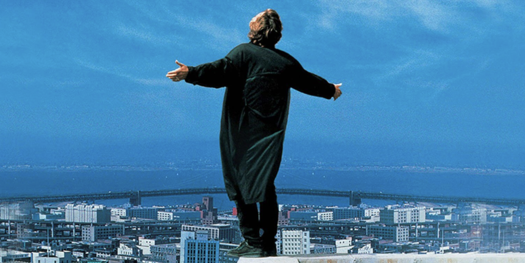 A man stands on the ledge of a building, holding his arms out in a Christ-adjacent pose. There's a bustling city in front of him with a large body of water in the distance.
