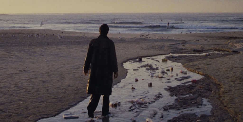 Richard Chamberlain wandering the beach. There is a river of water cutting through the beach, heading toward the Ocean.