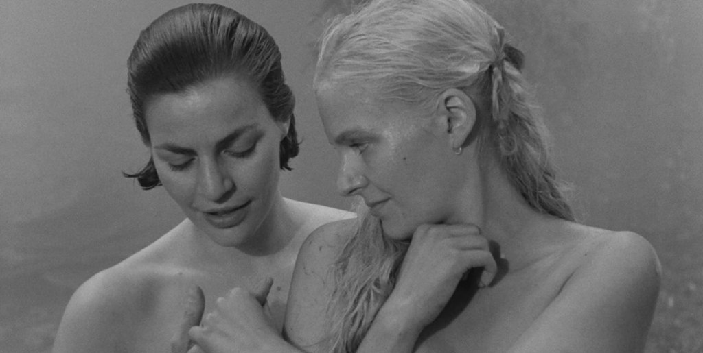 Two women, naked with wet hair, stand close together. The blonde woman is holding the dark haired woman's hand.