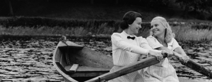 Two women, one with dark hair and one with light blonde hair, stare lovingly at each other while they row a boat on open water.