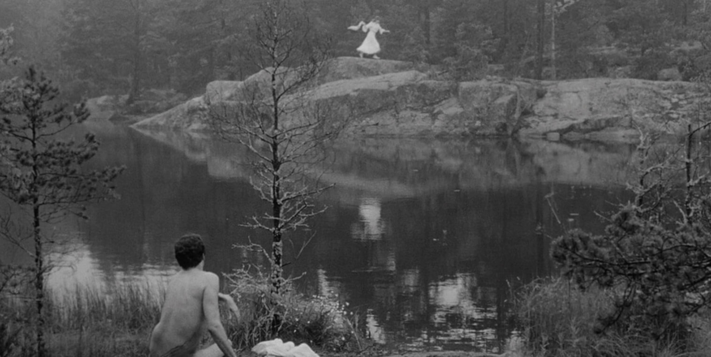A woman in a white dress stands on a bluff above a body of water. A naked person of undetermined gender sits with their back to the camera on the shore across from the bluff.