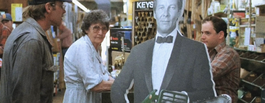 Allie Fox (Harrison Ford) is in a hardware store. He is standing to the right of the frame, staring at an old woman and an employee in a flannel. In between the woman and the employee is a black and white standee holding a Ninja chainsaw.