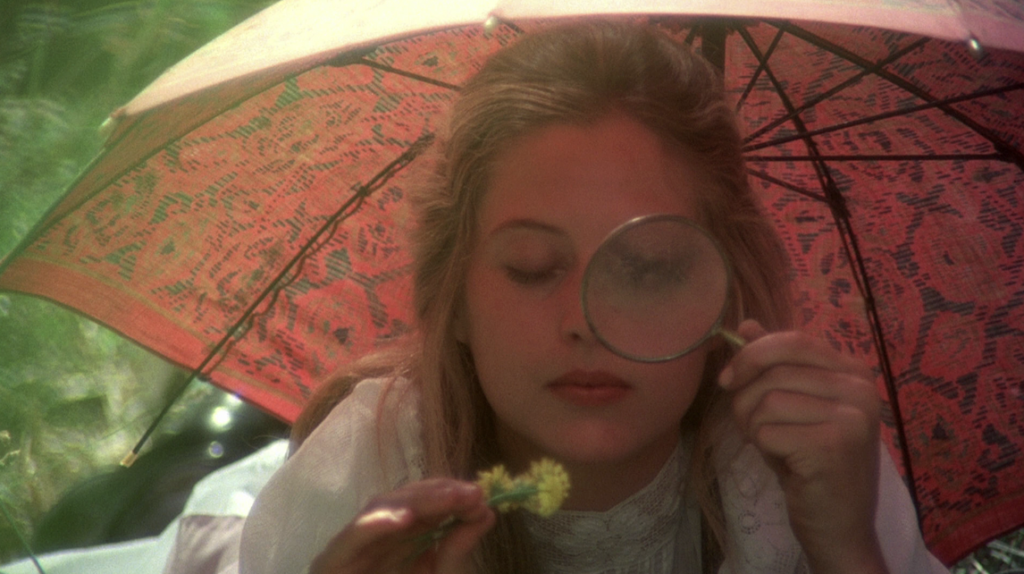 A young blonde woman examines a yellow flower with a large magnifying glass. She has a pink lace umbrella over her head.
