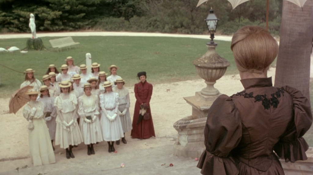 The headmistress, dressed in an awful brown gown with puffy sleeves, addresses a group of young women from the right of the image. The young women are standing below her in the left of the image, and are all wearing the same horrid white outfit with matching hats and gloves. There is a woman on the far left of the group holding an umbrella, and a woman on the right of the group dresssed in a red overcoat.