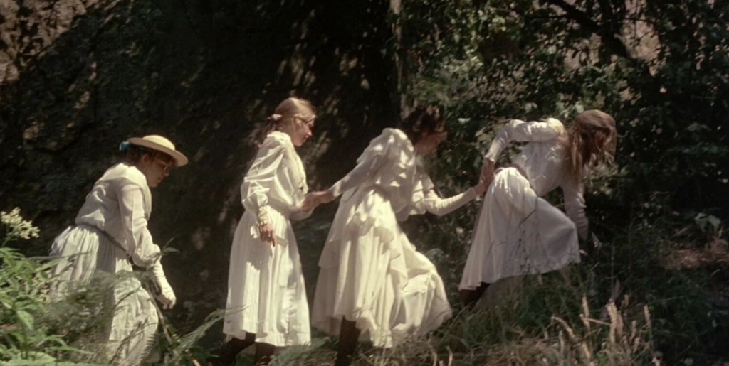 Four girls in white dresses scale a wooded hill; the one of the farthest left of the image is wearing a sun hat, the other three are holding hands and are not wearing any hats.