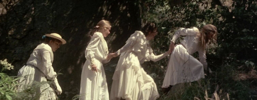 Four girls in white dresses scale a wooded hill; the one of the farthest left of the image is wearing a sun hat, the other three are holding hands and are not wearing any hats.