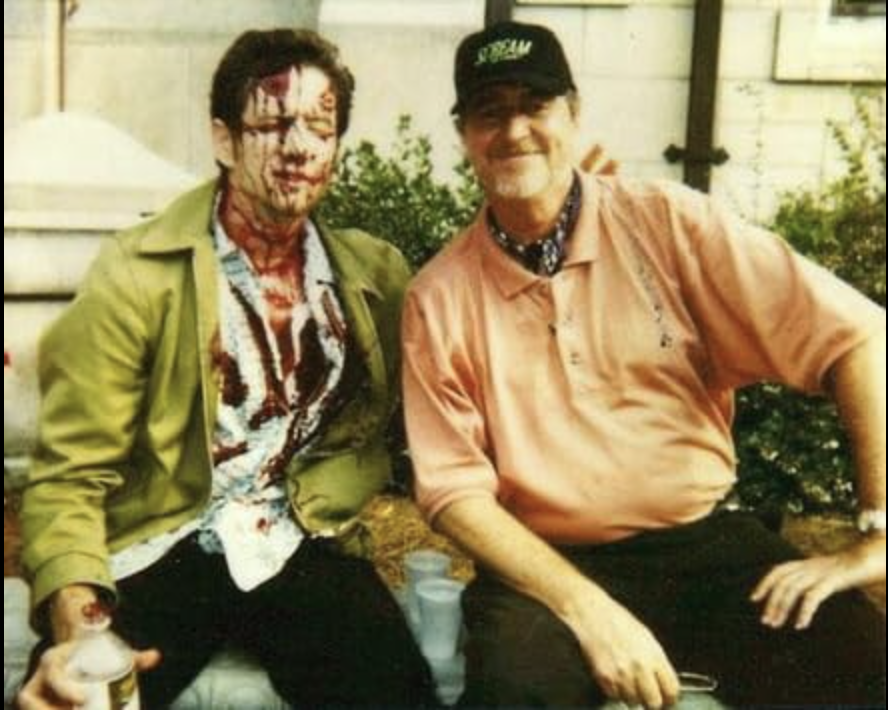 Jamie Kennedy (left) is sitting in a green jacket and a bloodied blue button-down. To his right is director Wes Craven, who is wearing a black hat and a delightful peach polo shirt.