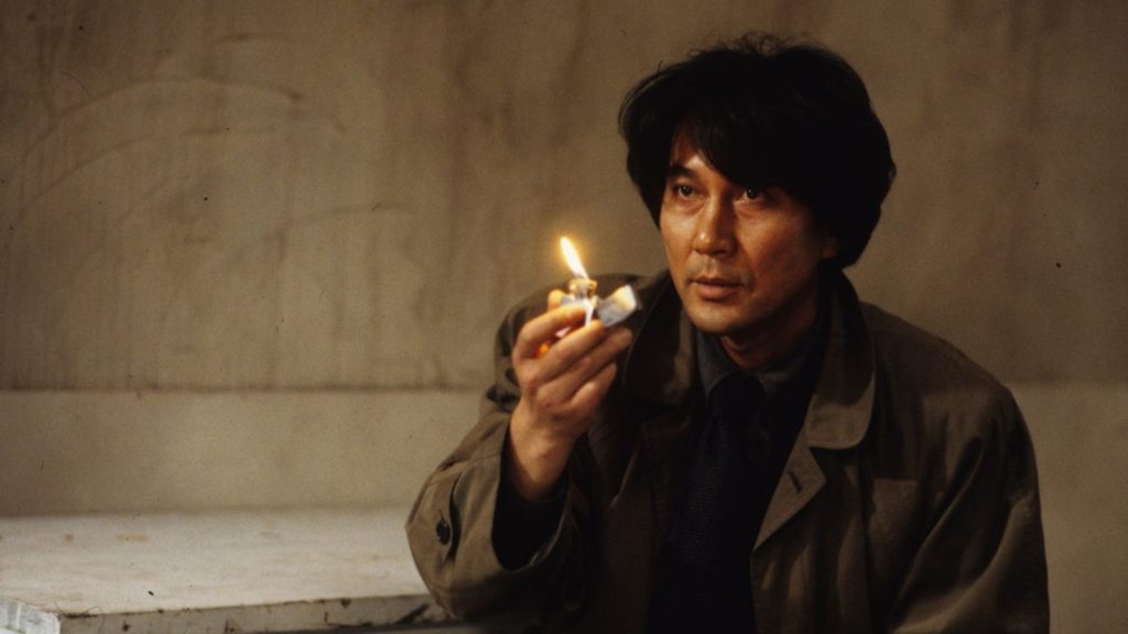 A middle-aged Asian man with tanned skin and medium long hair is sitting in a room with worn-out white walls. He is holding a lit lighter in front of his dark brown Trenchcoat, looking off camera