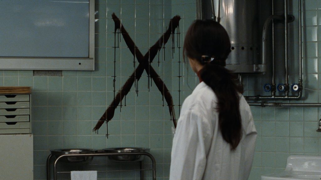 A woman with long dark hair is facing away from the camera, examining a large, x-shaped cross that is painted on a mint-tiled wall in what appears to be a medical examination room.