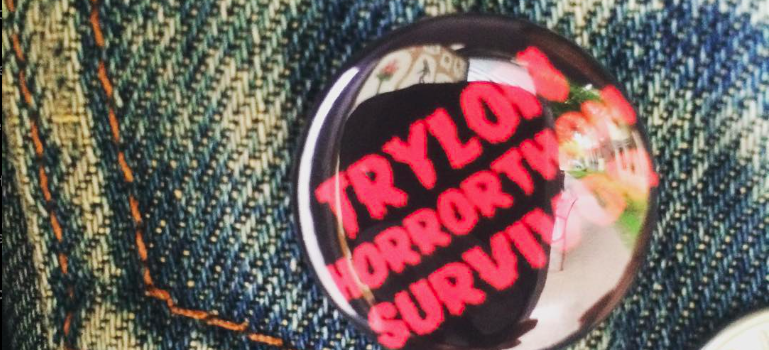 Closeup of a small, shiny black button with a red bold font that reads "Trylon Horrorthon Survivor" pinned onto the top of a pocket of a light blue denim jacket.