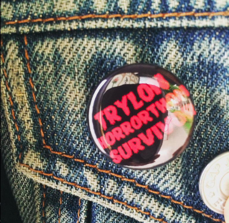 Closeup of a small, shiny black button with a red bold font that reads "Trylon Horrorthon Survivor" pinned onto the top of a pocket of a light blue denim jacket. 