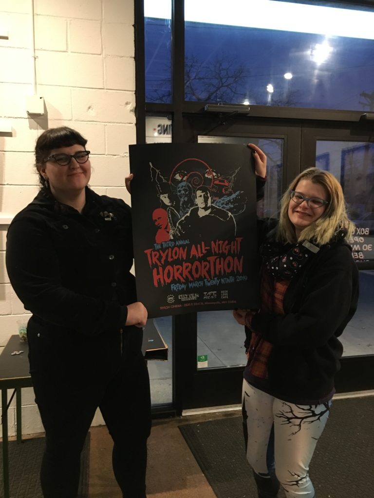 Two very tired yet happy looking individuals holding up a poster with drawings of horror movie characters and script in red bold font that reads "Trylon All-Night Horrorthon"