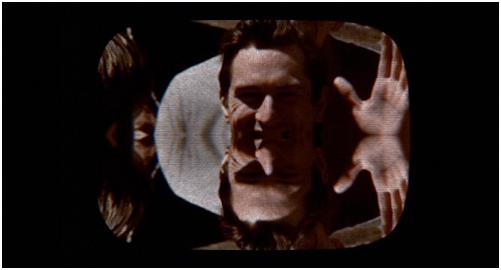 A white man waving at the camera, with the image cut off horizontally and mirrored at the middle