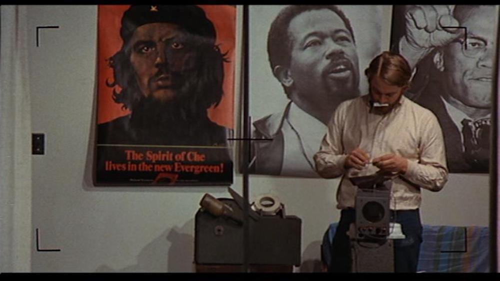 A white man standing behind an old film projector on a table, with posters of Che Guevara, Eldridge Cleaver, and Malcolm X on the wall behind him