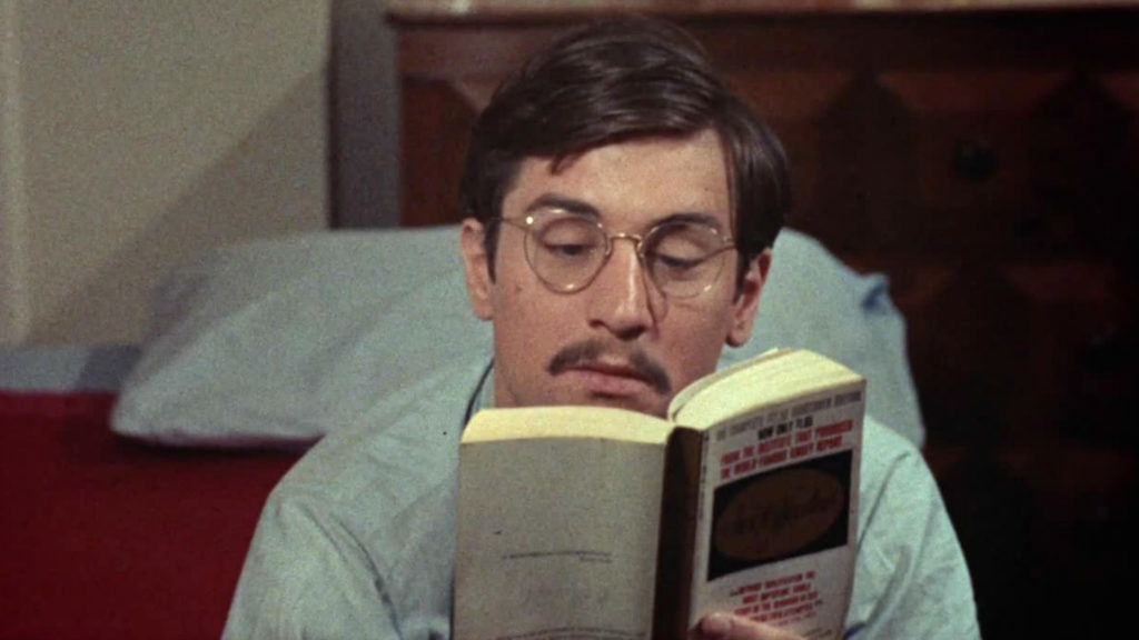 Robert De Niro plays Jon Rubin in Greetings. Rubin is a light-skinned man with medium-long brown hair, eyeglasses, and a mustache. Sitting on a bed, he is reading a book.